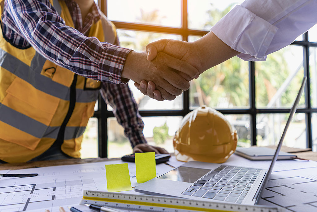 Two contractors shaking hands over architectural plans and a laptop during a luxury home renovation meeting